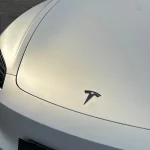 Tesla logo close-up hood shot MOZX White Iridescent Gold PET vinyl wrap, soft golden light reflection on white PET surface
