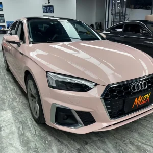 Front three-quarter shot Audi A5 Sportback wrapped with MOZX Smart Shell Pink high gloss PET vinyl wrap, creamy pastel pink paint-like finish in professional car wrapping shop Title